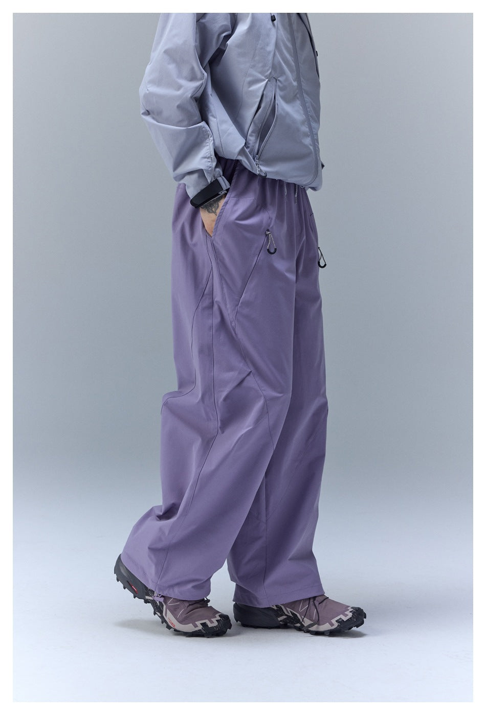Side view of a person wearing loose lavender pants with zip pockets, a light gray jacket, and trail shoes.