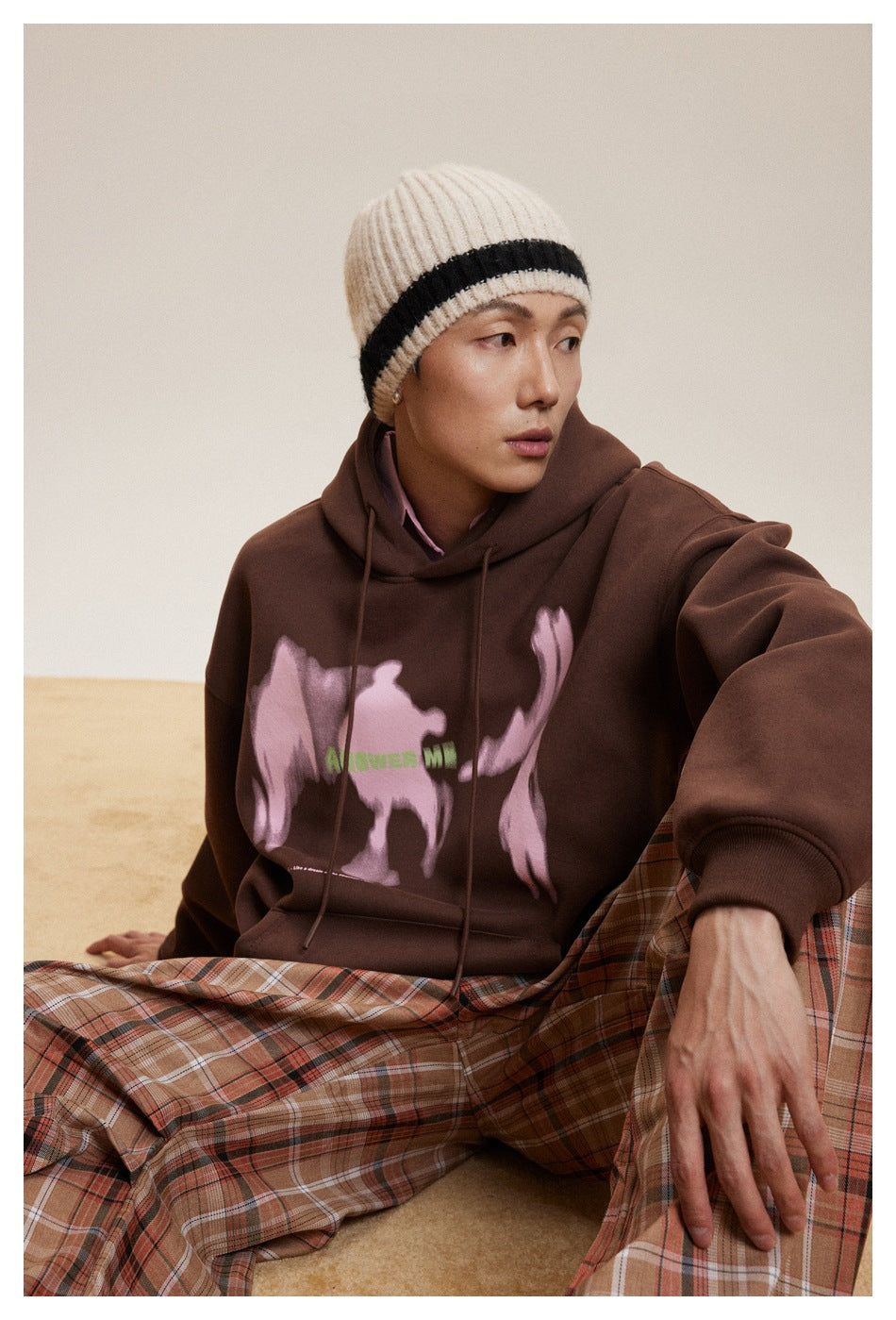 Seated model in a dark brown hoodie with a pink abstract graphic and rust plaid trousers.