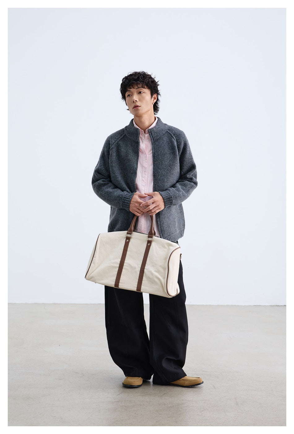 Model wearing an open charcoal gray zip-front knit jacket over a pale pink shirt, holding a cream duffel bag.