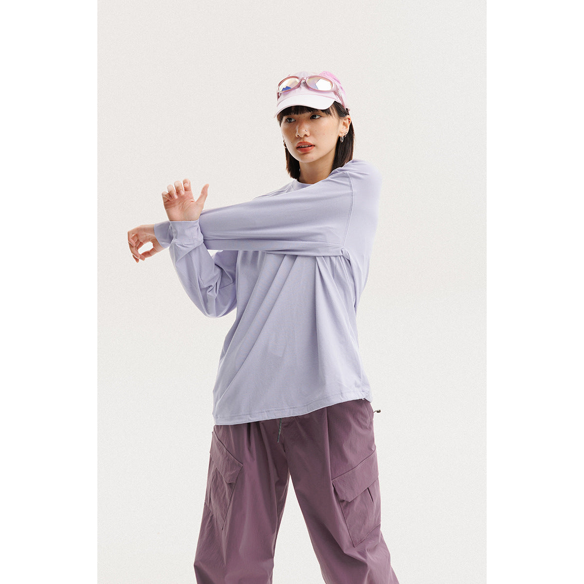 Model wearing a pale lavender long-sleeve shirt and mauve cargo pants, stretching one sleeve; pink cap and goggles.