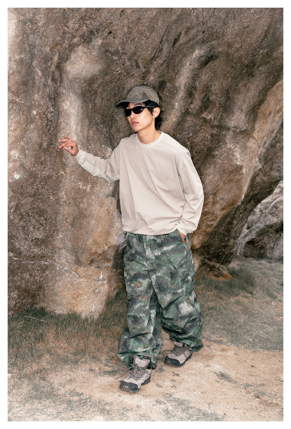 Model wearing a loose cream long-sleeve shirt and wide green camouflage cargo pants, posing beside a rocky wall.