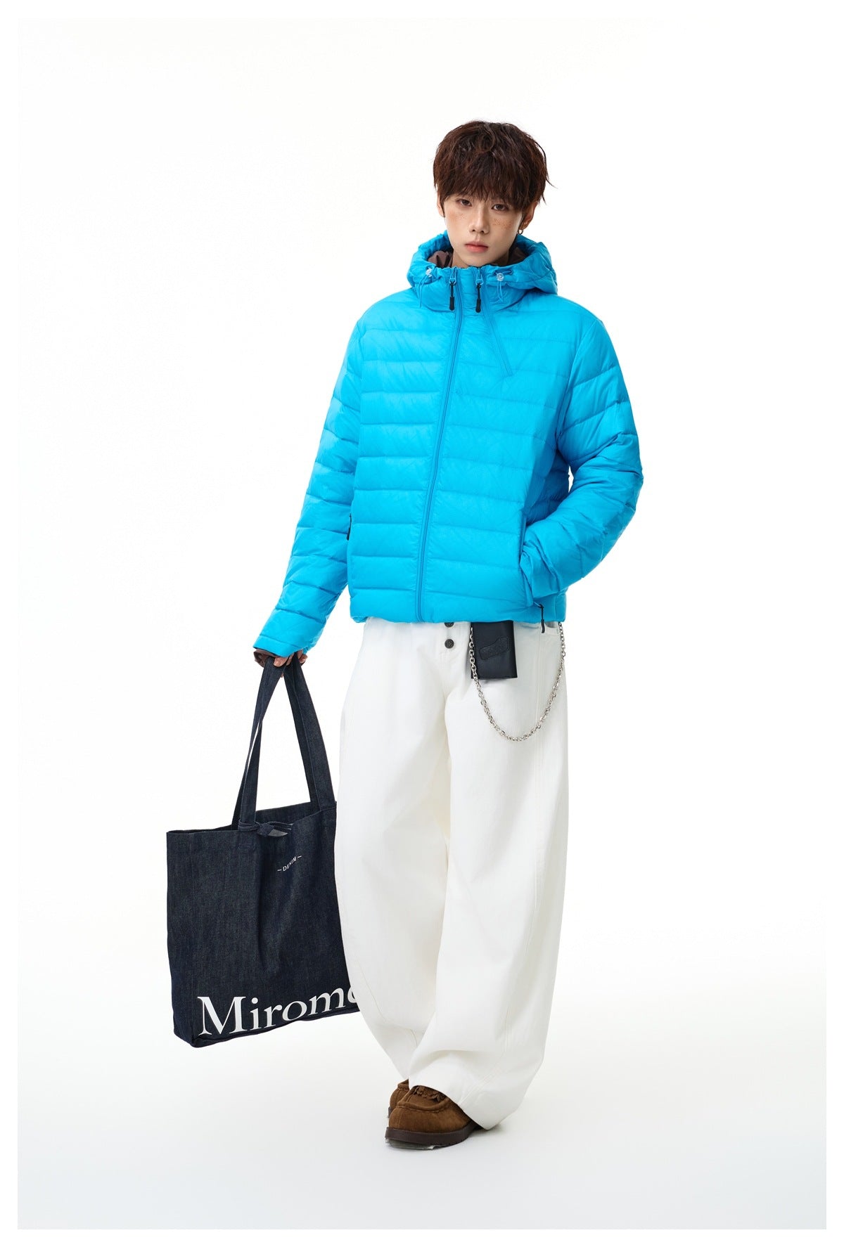 Full-length model wearing a bright cyan quilted hooded jacket and loose white trousers, carrying a dark tote bag.