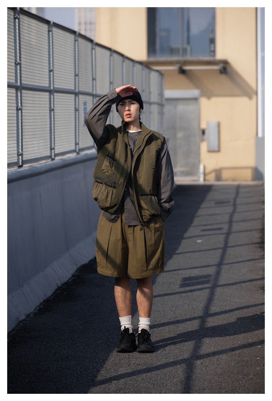 Model wearing an olive utility vest over a gray shirt with matching loose olive shorts.