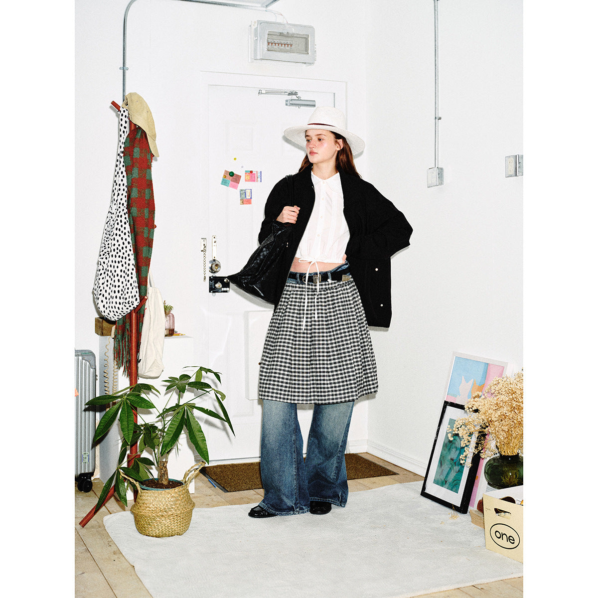 Model wearing an oversized black jacket over a white blouse and checked skirt with wide-leg jeans and a white hat.