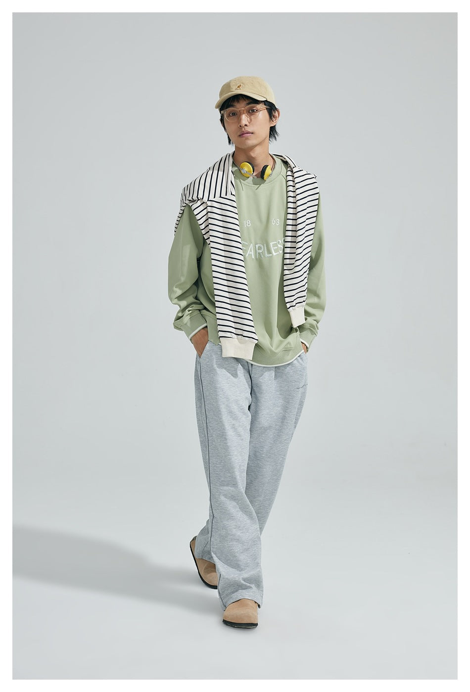 Model wearing a light green sweatshirt with a striped sweater over the shoulders, gray wide-leg pants and a tan cap