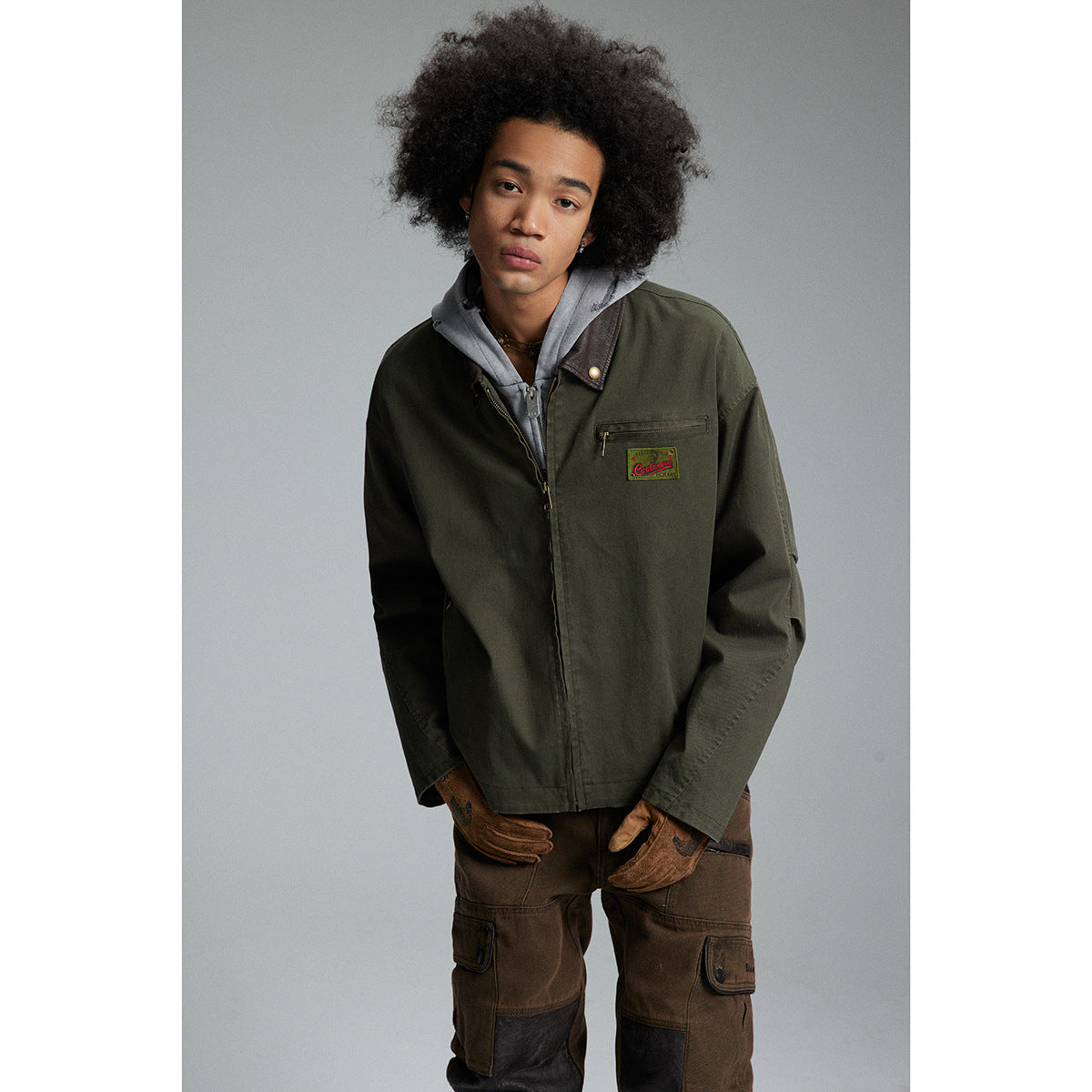 Model in an olive work jacket with a zip chest pocket and patch, layered over a gray hoodie.