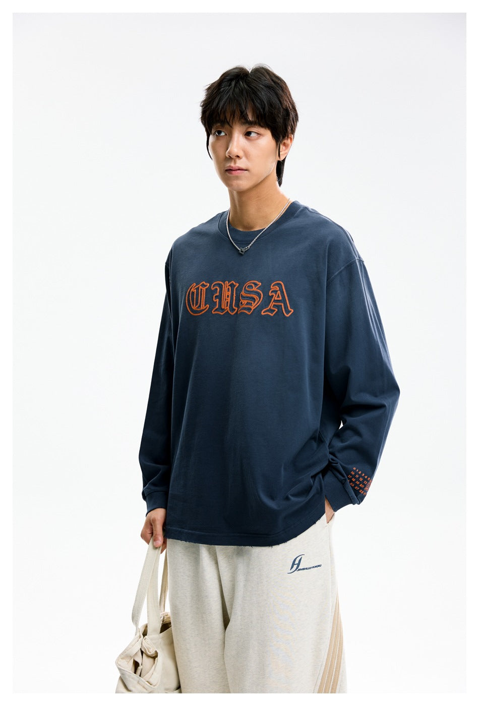 Model wearing a navy long-sleeve shirt with orange USA lettering and cream pants, carrying a canvas tote.