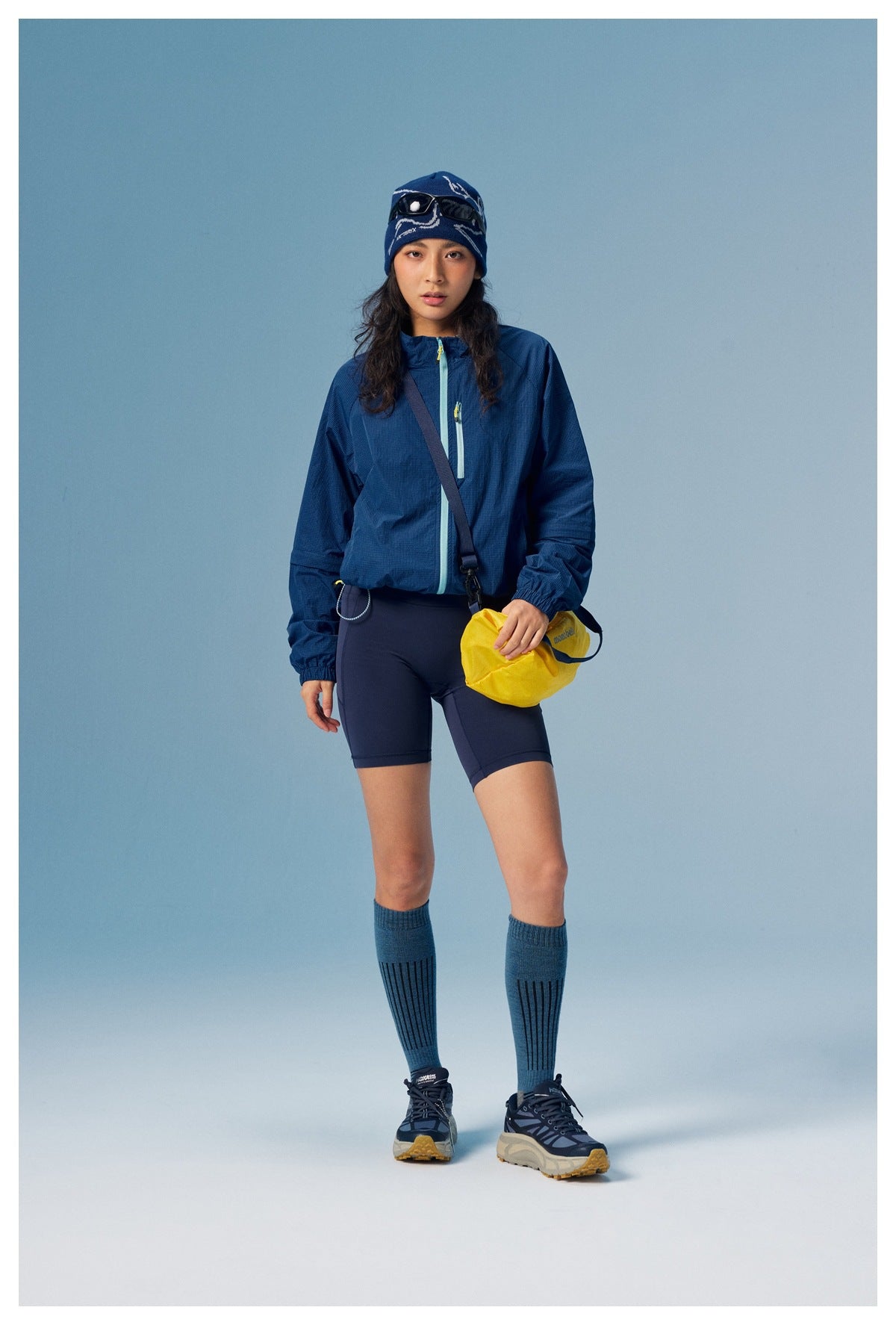 Front view of a model wearing a navy zip-front jacket and dark shorts with blue knee socks and a yellow shoulder bag.