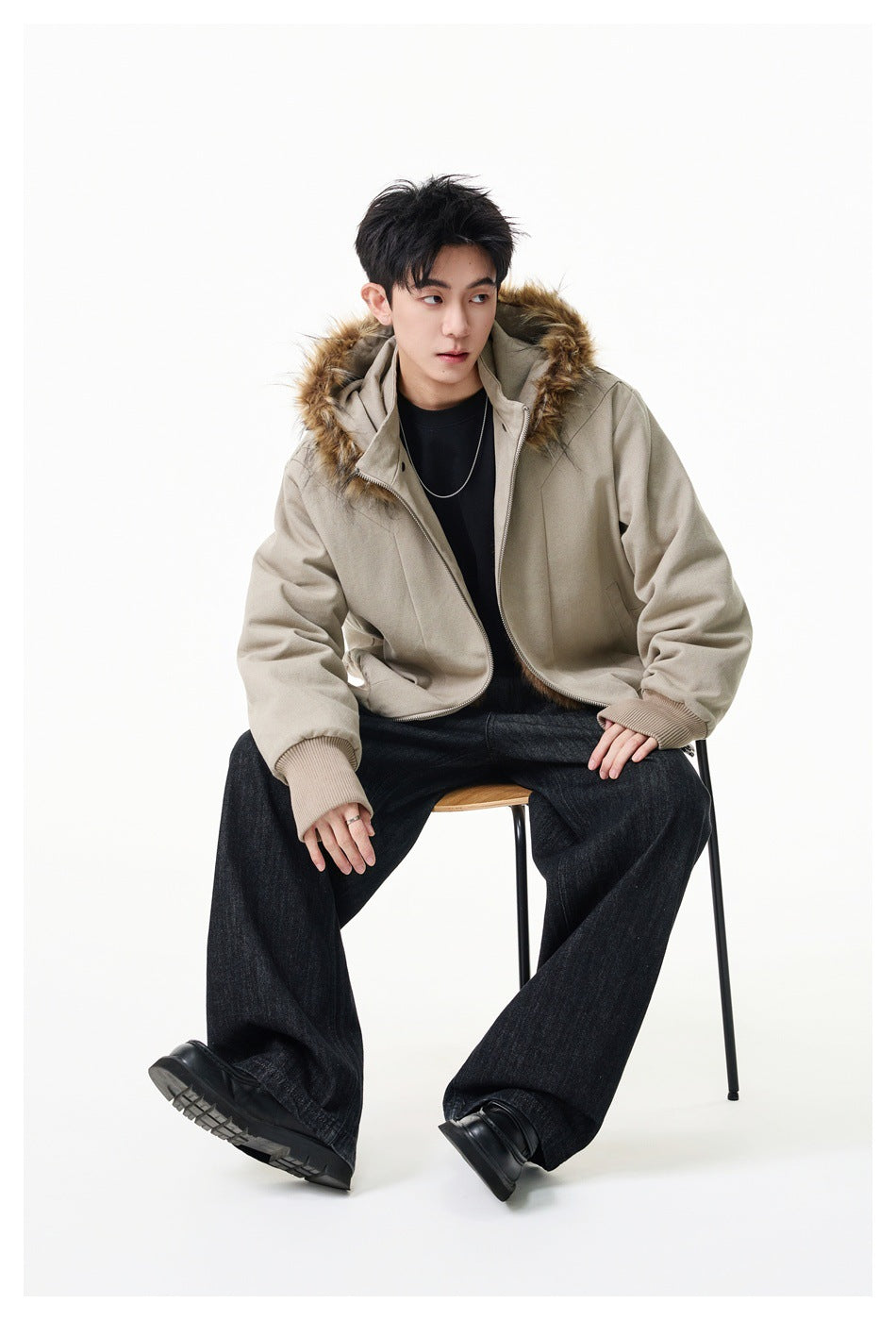 Seated model wearing an open light beige hooded jacket with tan fur trim and ribbed cuffs, paired with dark wide-leg pants.