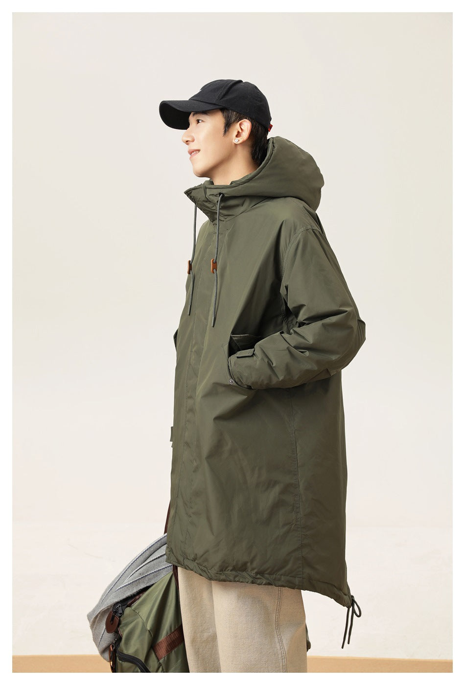 Model in a long olive hooded parka with drawstrings and a black cap, shown from the side.