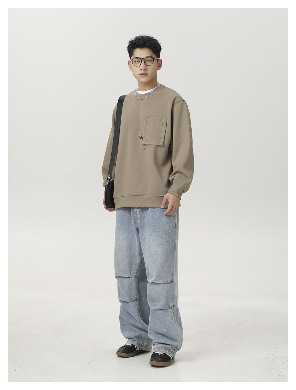 Man wearing a taupe crewneck sweatshirt with a large chest pocket and loose light-wash jeans, shown full-length.