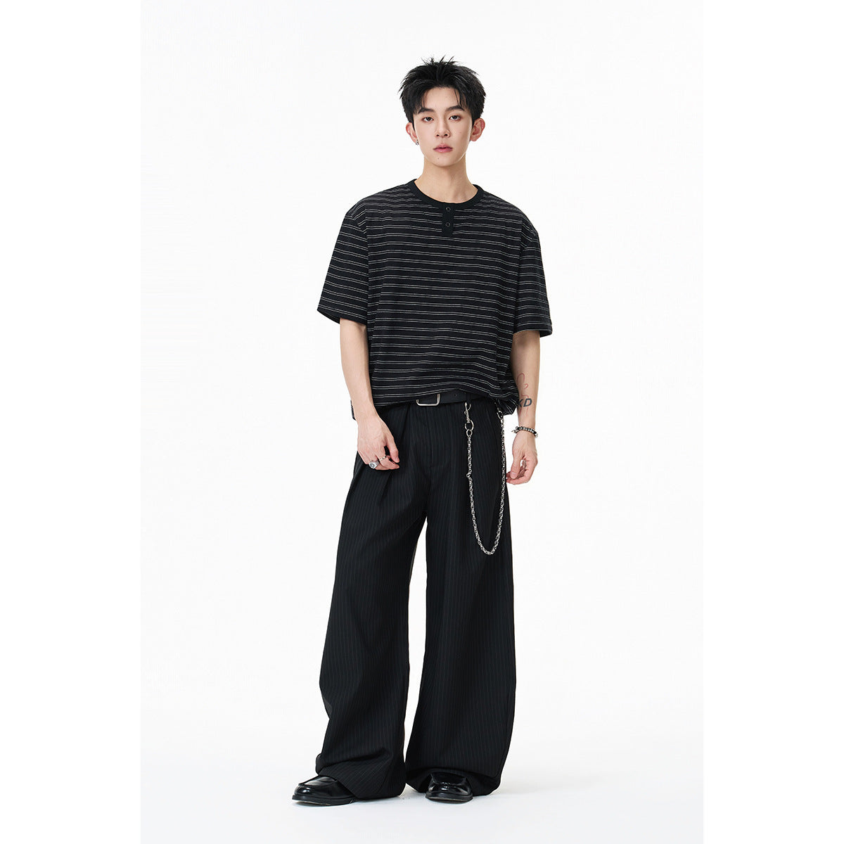 Model wearing a black short-sleeve Henley with thin gray stripes and loose black trousers.