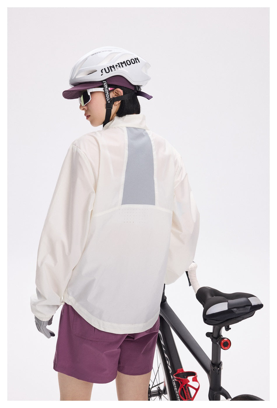 Rear view of a cyclist wearing an ivory jacket with a gray back panel and plum shorts beside a road bike.