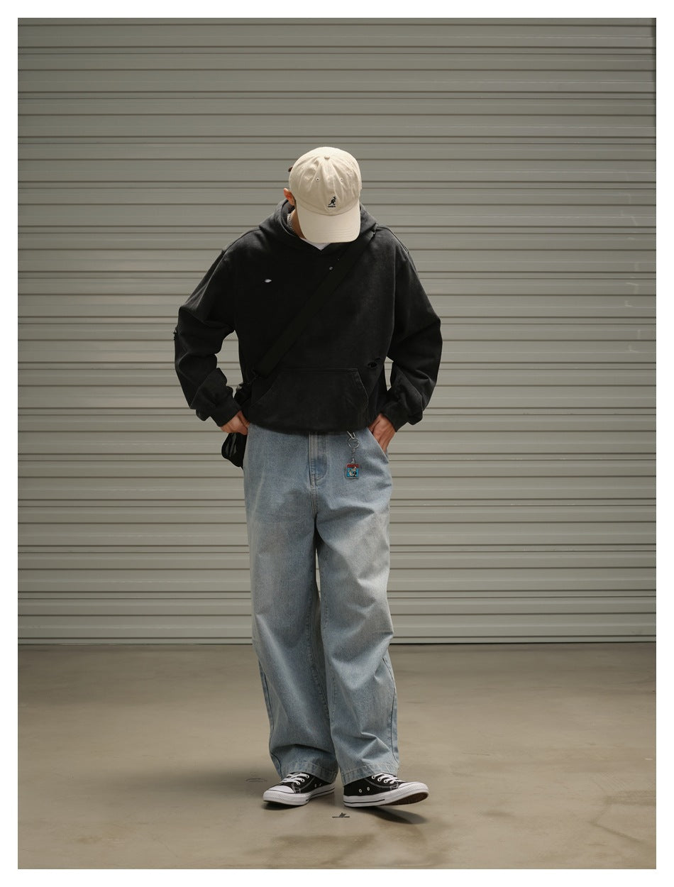Model looking down in a black distressed hoodie, loose blue jeans and cream cap, carrying a black crossbody bag.