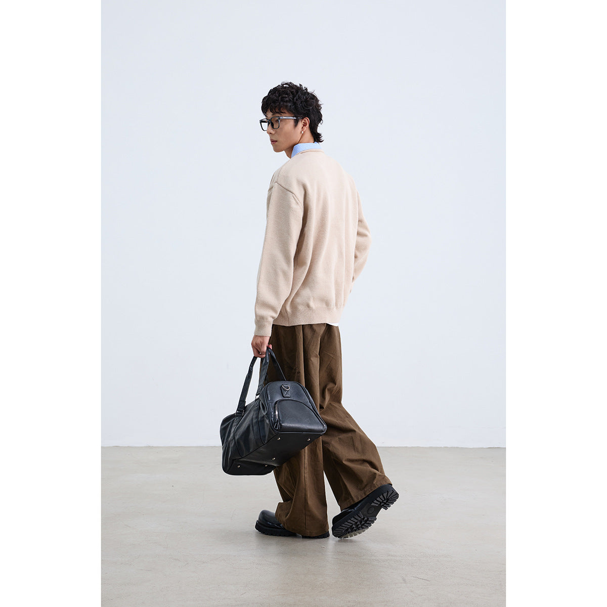 Model in a beige sweater, shown from the back and side with brown trousers and a black bag.