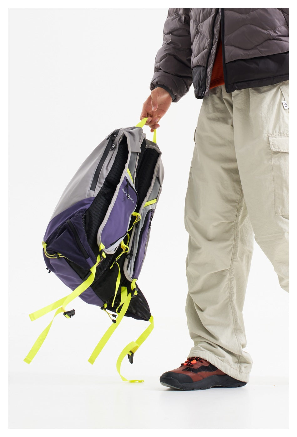 A person holds a gray and purple backpack by its neon yellow handle, showing the padded mesh back and loose straps.