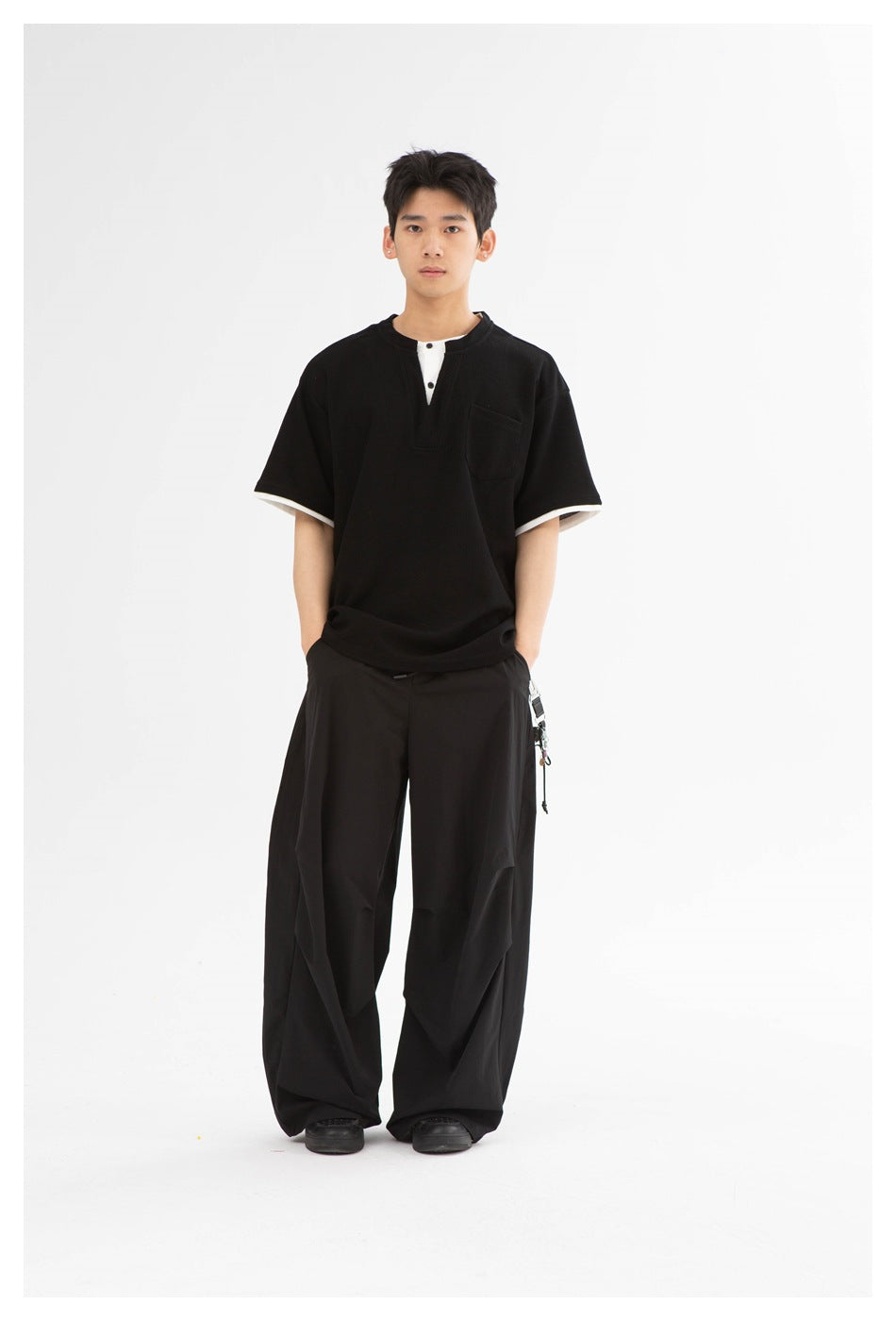 Full-length front view of a man wearing a black short-sleeve henley with white trim and loose black trousers.