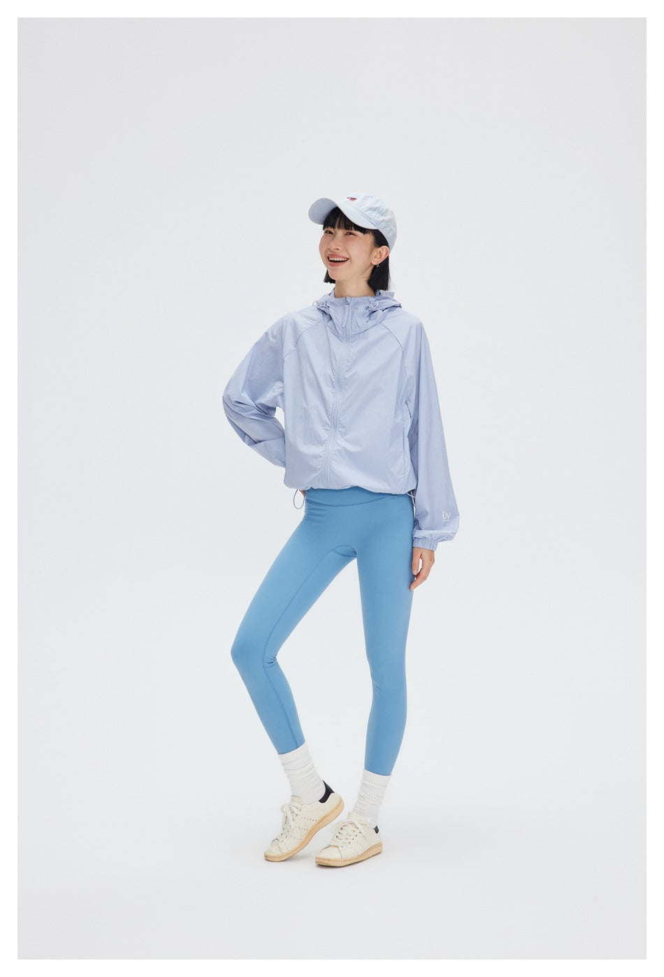 Full-length view of a woman wearing a pale blue hooded jacket, blue leggings, and a white cap.