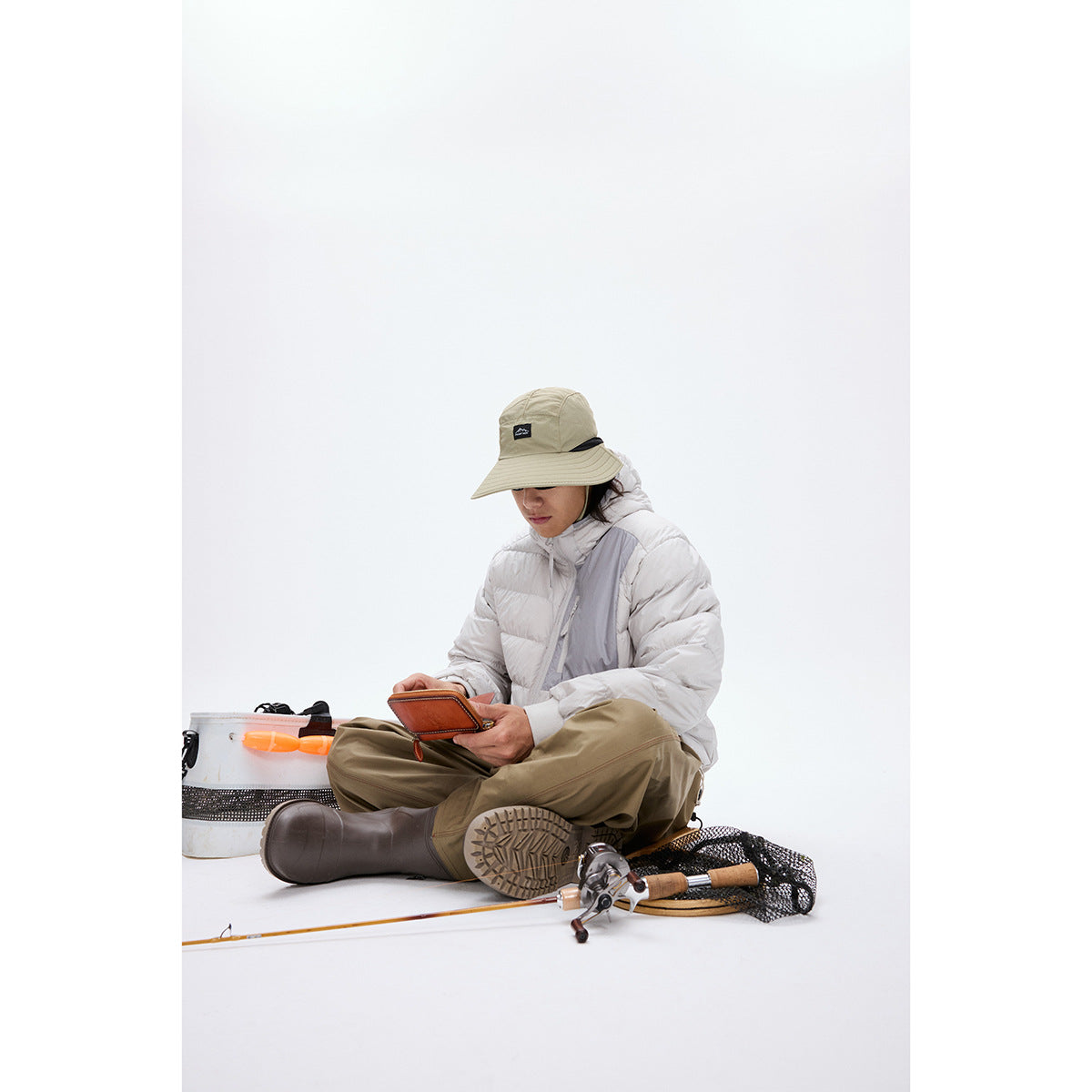 A seated model wears a light gray hooded puffer jacket and khaki trousers among fishing gear.