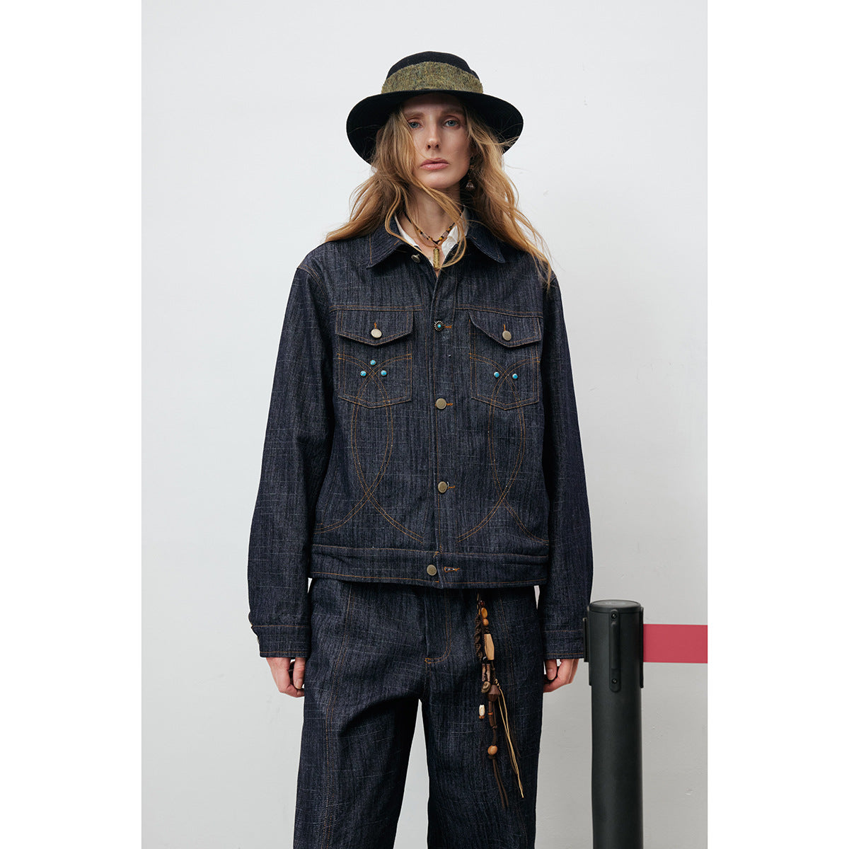 Model wearing a dark indigo button-front jacket with decorative stitching and matching trousers, styled with a wide-brim hat.