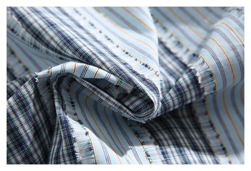Close-up of draped light blue shirt fabric showing dark plaid bands, narrow stripes, and fringed seams.