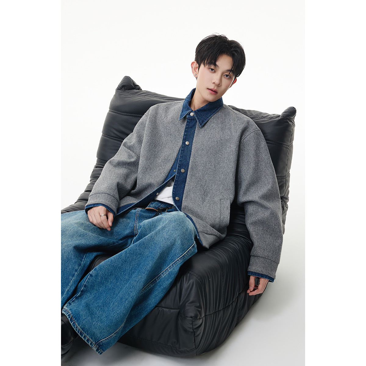 Seated model wearing an oversized gray jacket with a blue denim collar and placket, paired with blue jeans.