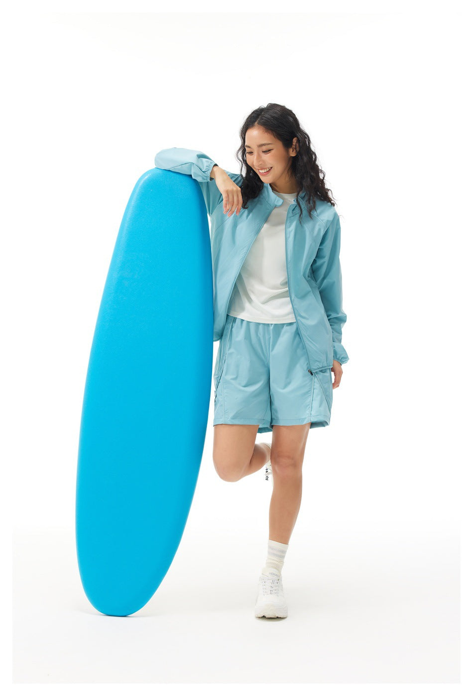 Model wearing a light blue zip jacket and matching shorts beside a large blue board.