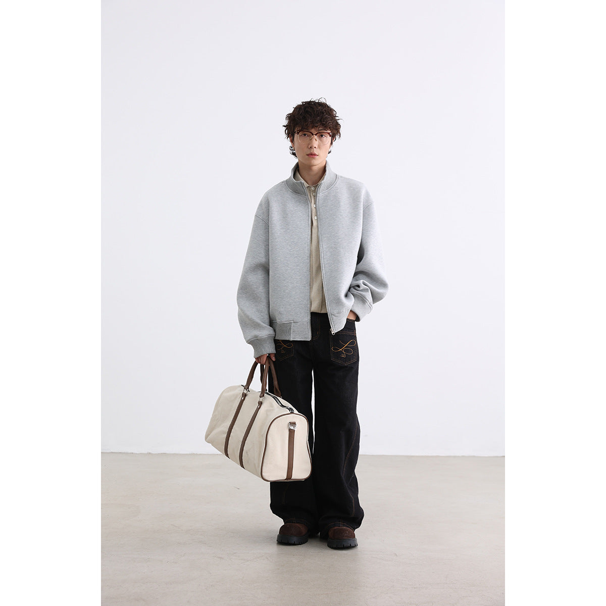 Full-length model wearing a light gray zip jacket with dark jeans and carrying a cream duffel bag.