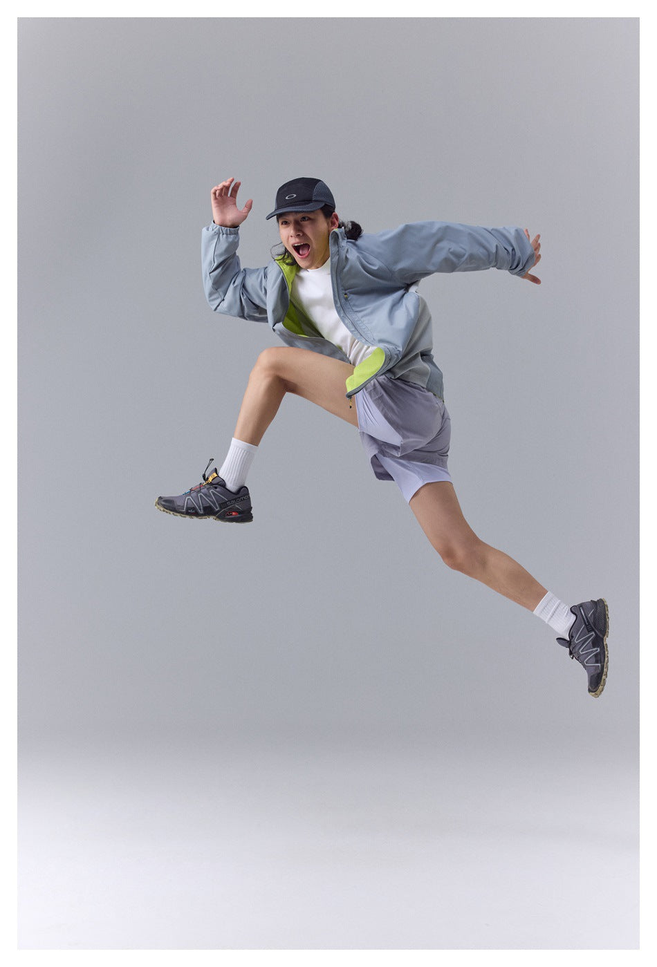 Model leaping in a blue-gray outdoor jacket with lime lining, a white shirt, pale shorts, and a dark cap.
