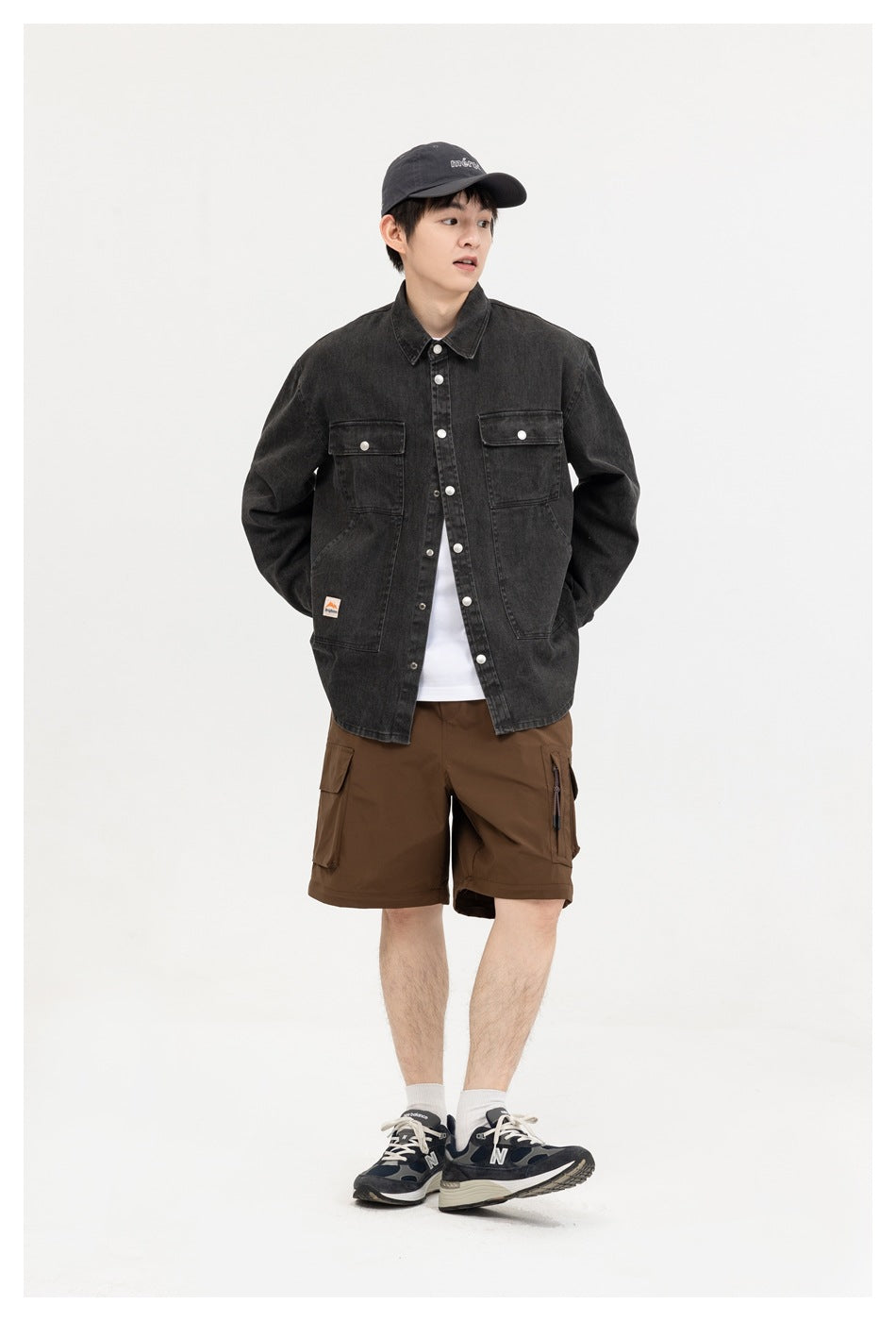 Model wearing the washed black button-front shirt open over a white tee with brown shorts, cap and navy sneakers
