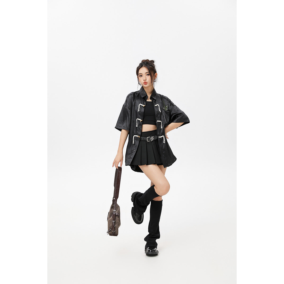 Model wearing an open black short-sleeve knot-front shirt over a black crop top and pleated mini skirt.