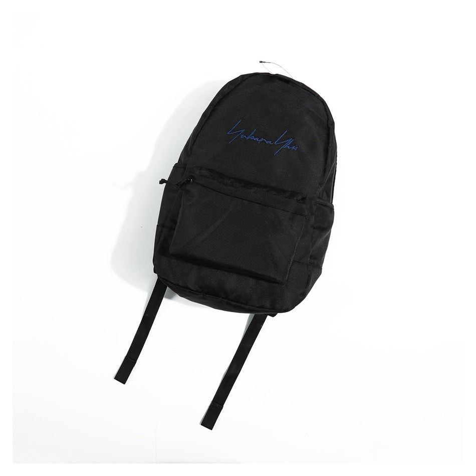 Black backpack flat lay with blue embroidered script and front pocket, shown at an angle on white