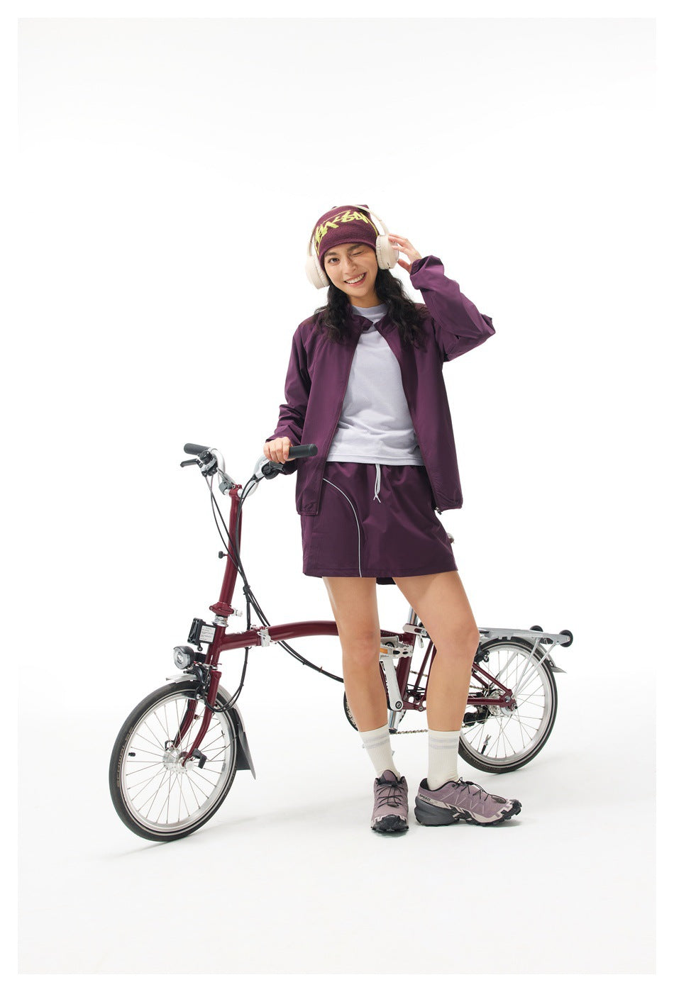 Model wearing a plum windbreaker and matching shorts beside a bicycle; yellow graphic on her plum headband.