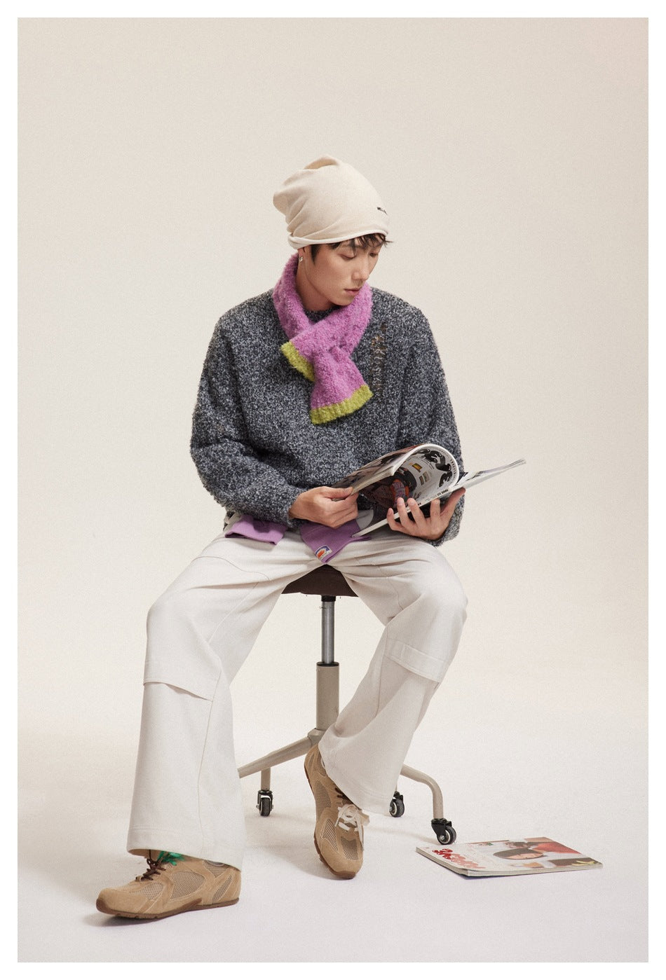 Model seated in a charcoal marled sweater, cream trousers, beige beanie, and pink scarf.