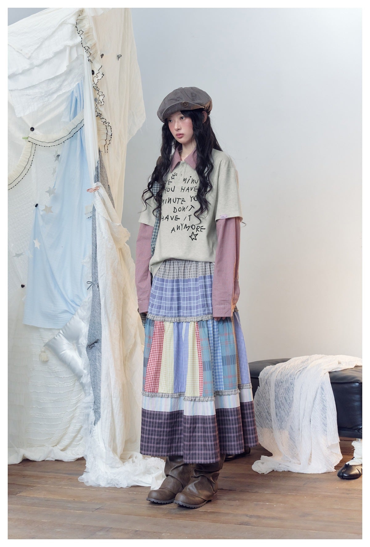 Model in a pale gray graphic tee layered over a pink checked shirt and a long tiered patchwork skirt.