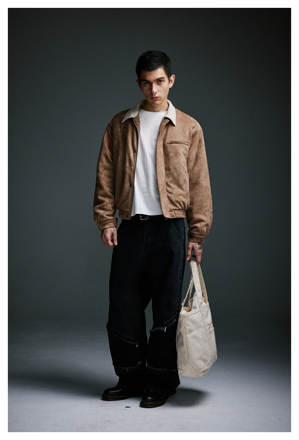Model standing in a khaki suede-look jacket with cream collar over a white tee, black jeans and boots, holding a tote