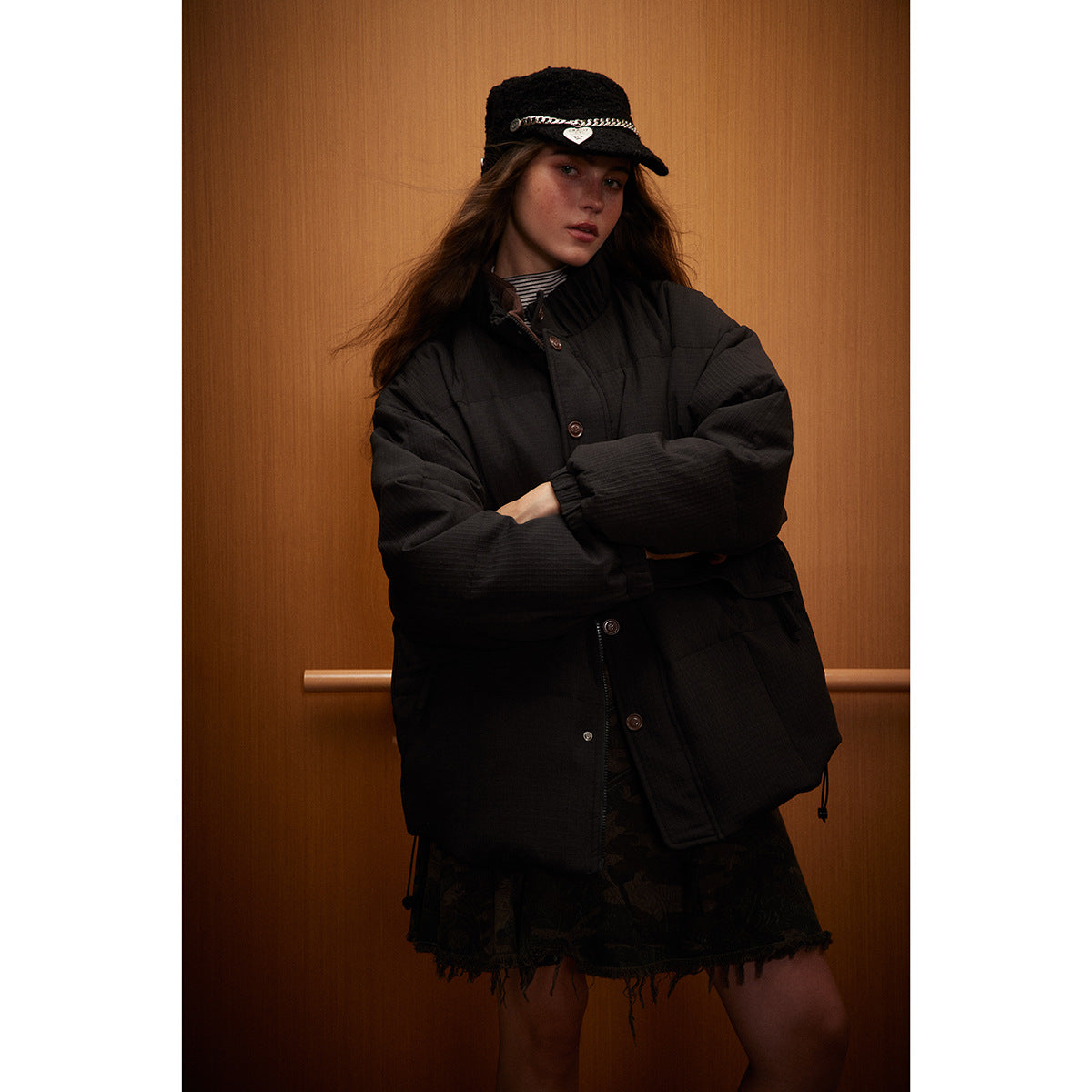 Model wearing a black padded button-front jacket and black cap with a silver chain and heart charm.