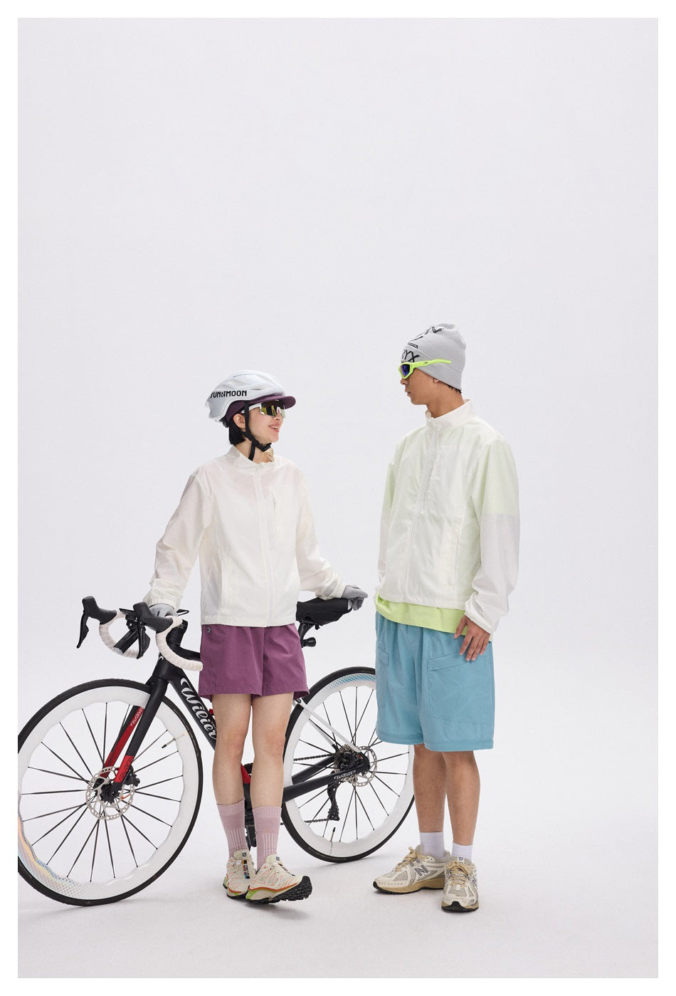 Two cyclists pose with road bikes, wearing light jackets and plum or blue shorts.