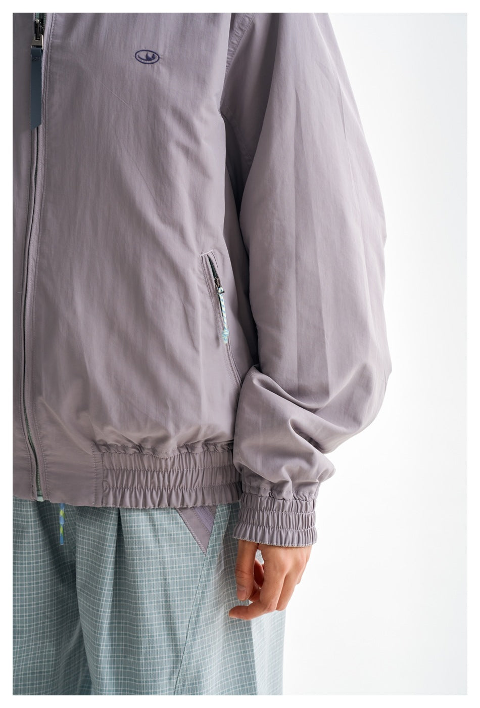 Close-up of the lavender-gray jacket's chest emblem, zip pocket, front zipper, gathered hem and cuff.