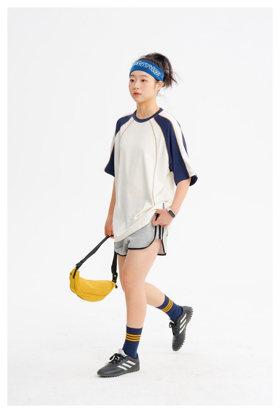 Model wearing a cream and navy raglan tee with gray shorts, carrying a yellow waist bag.