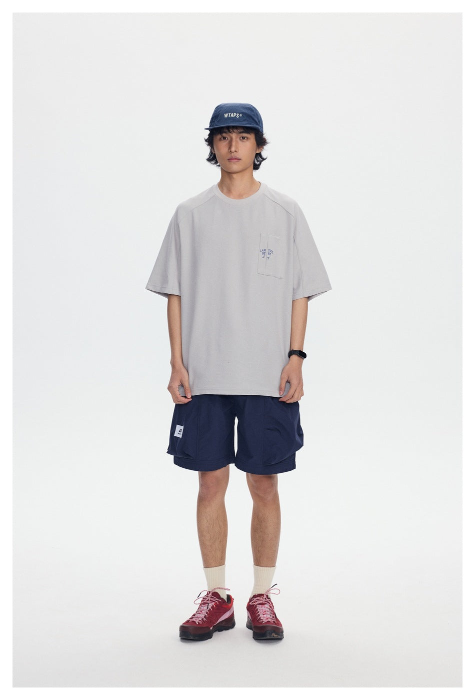Model wearing light gray relaxed crew-neck T-shirt with chest pocket, navy shorts, cap and red sneakers, front view