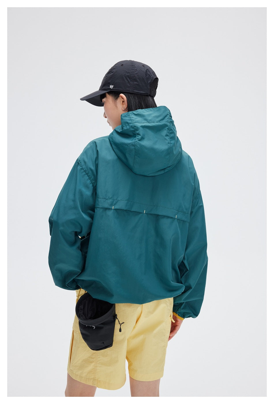 Back view of a model wearing a teal hooded jacket and pale yellow shorts.