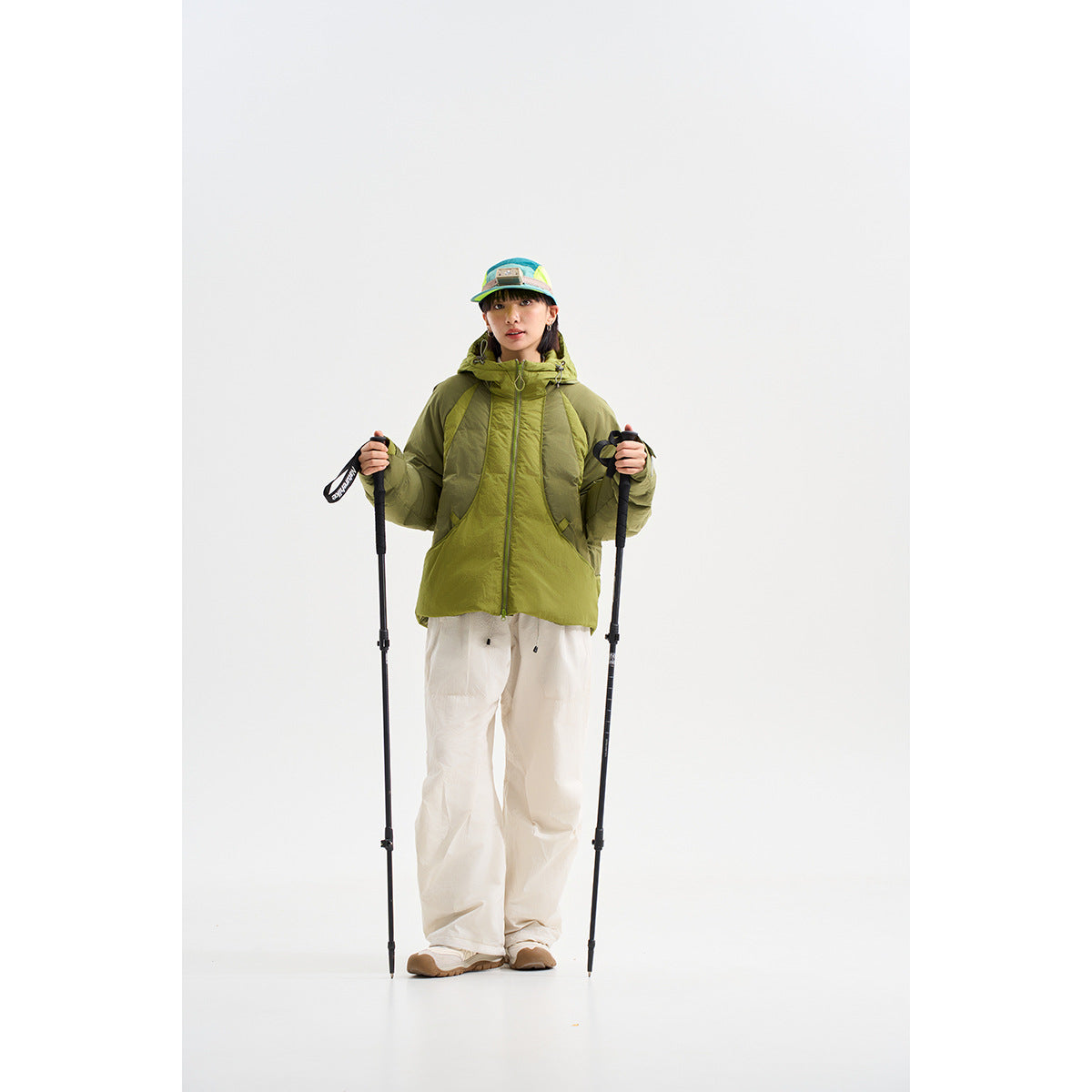 Model wears an olive-green hooded jacket with white pants and holds two trekking poles.