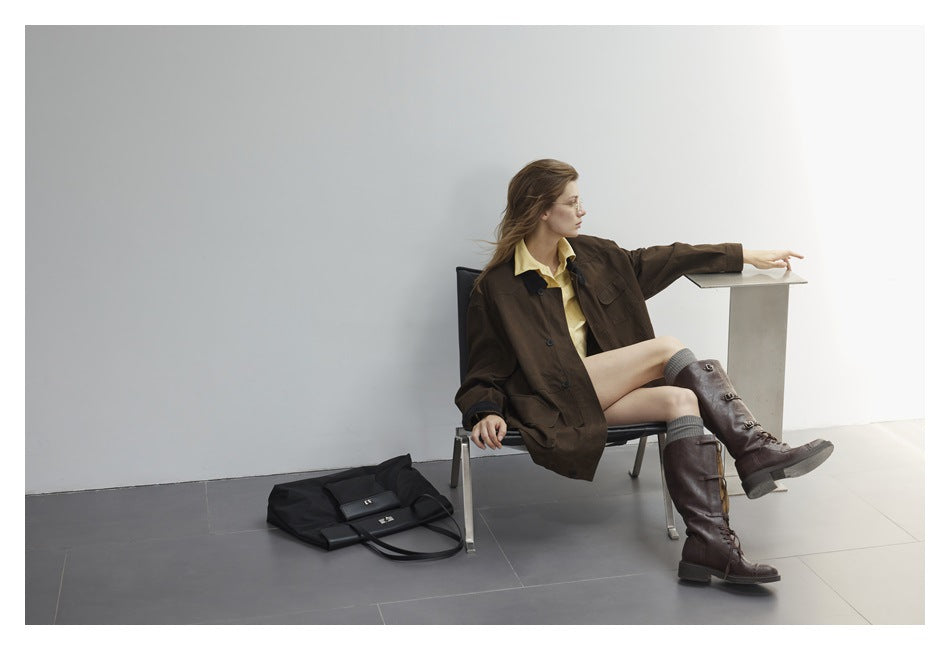Wide view of a seated model in a dark brown oversized jacket, yellow shirt, and tall brown boots.