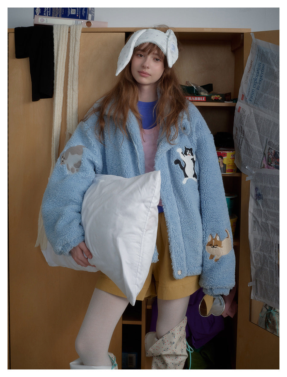 Model wearing an oversized light-blue plush jacket with animal appliqués, shown from the front while holding a white pillow.