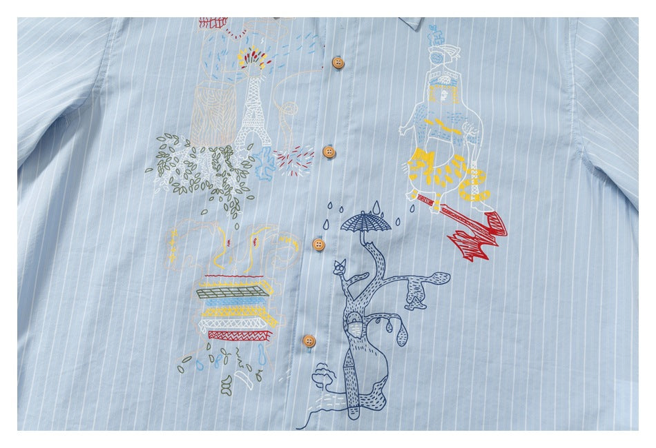 Close-up of colorful illustrated embroidery and tan buttons on the front of a light blue pinstriped shirt.