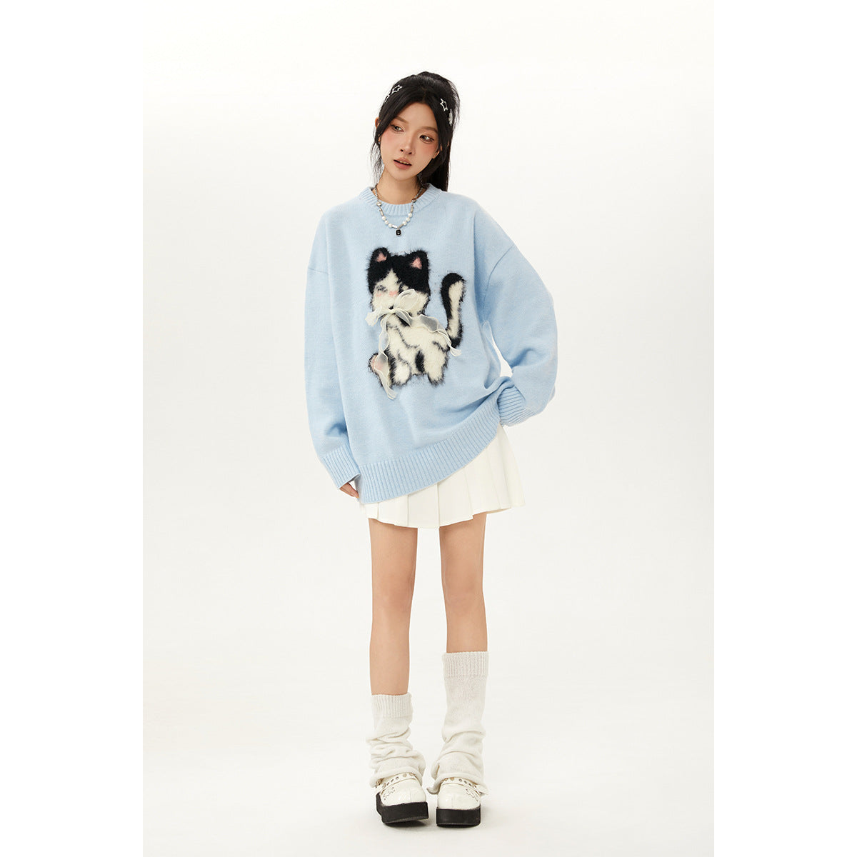 Model wearing an oversized light blue sweater with a fluffy black-and-white cat applique and a white pleated skirt.