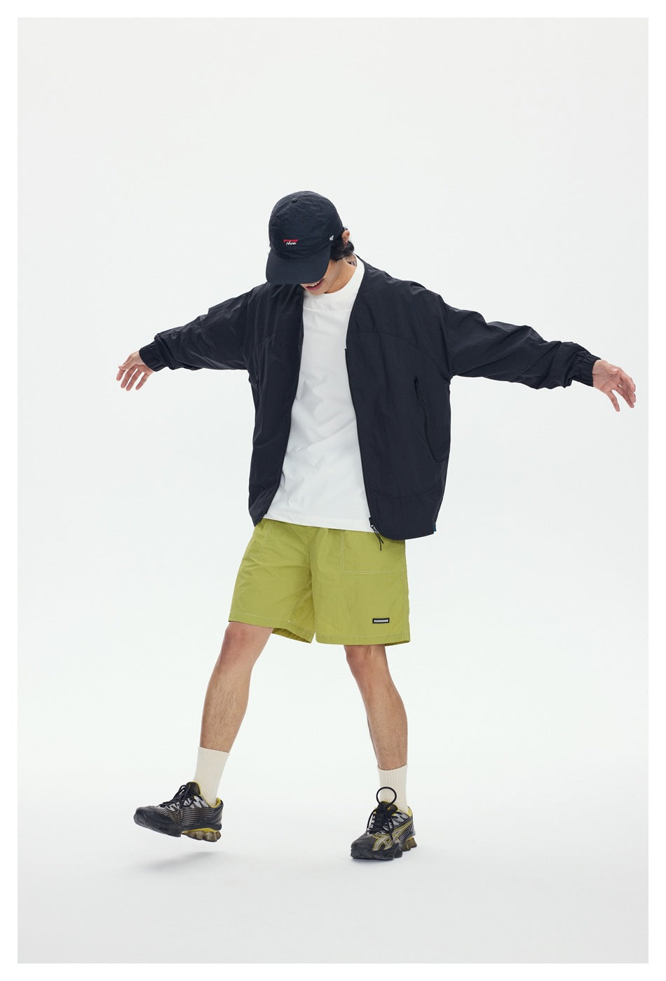 Model wearing a black open jacket, white shirt and lime green shorts with arms outstretched.