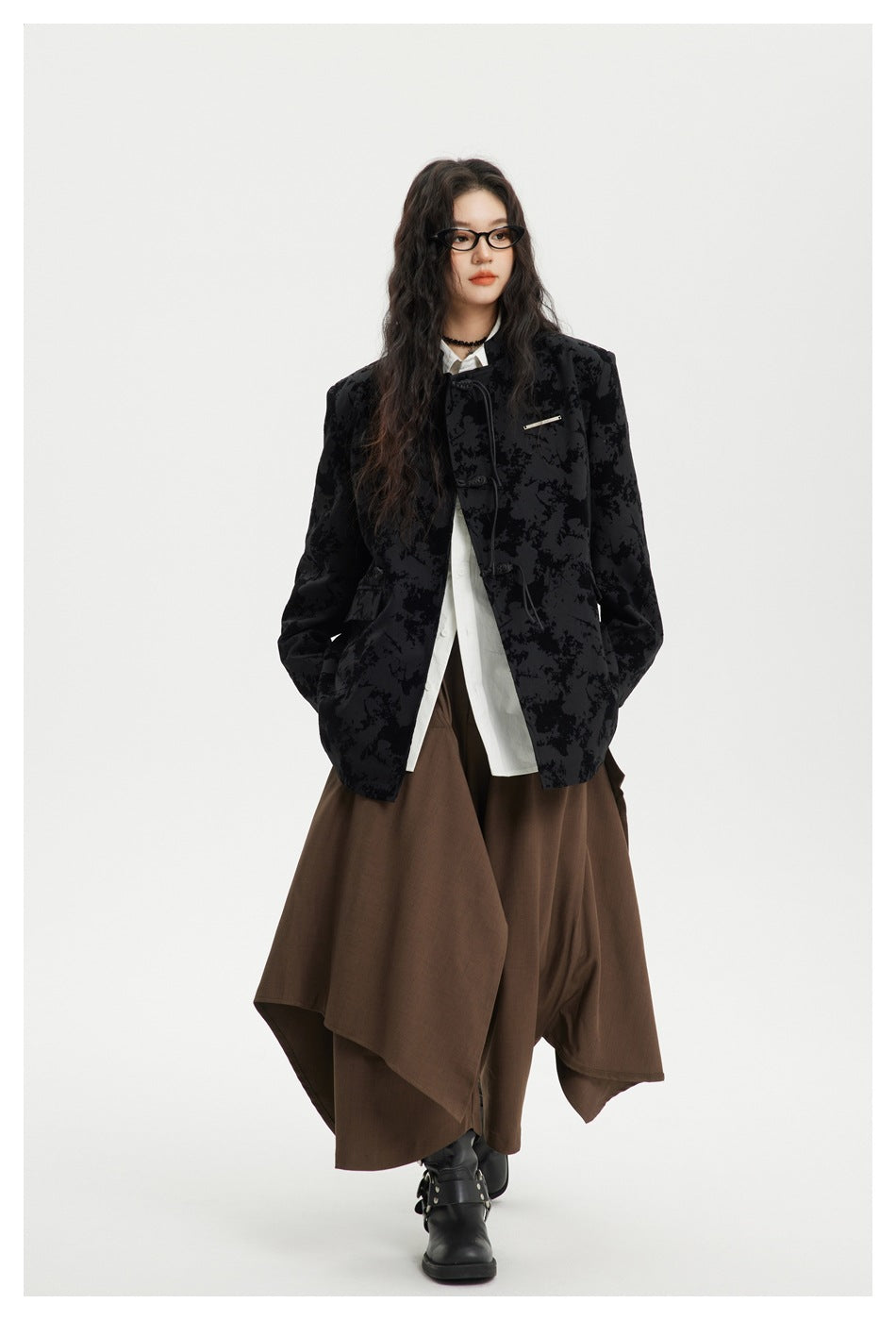 Model walking in a black abstract-patterned toggle-front jacket, white shirt, brown draped wide-leg trousers and black boots.