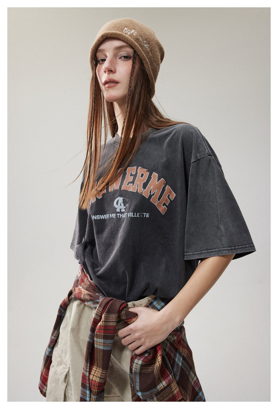 Model in a charcoal oversized graphic T-shirt and tan beanie, with a plaid shirt tied around her waist.